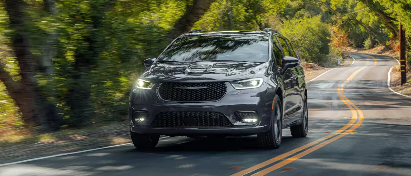 2026 Chrysler Pacifica black minivan driving on a tree-lined road in front three-quarter view